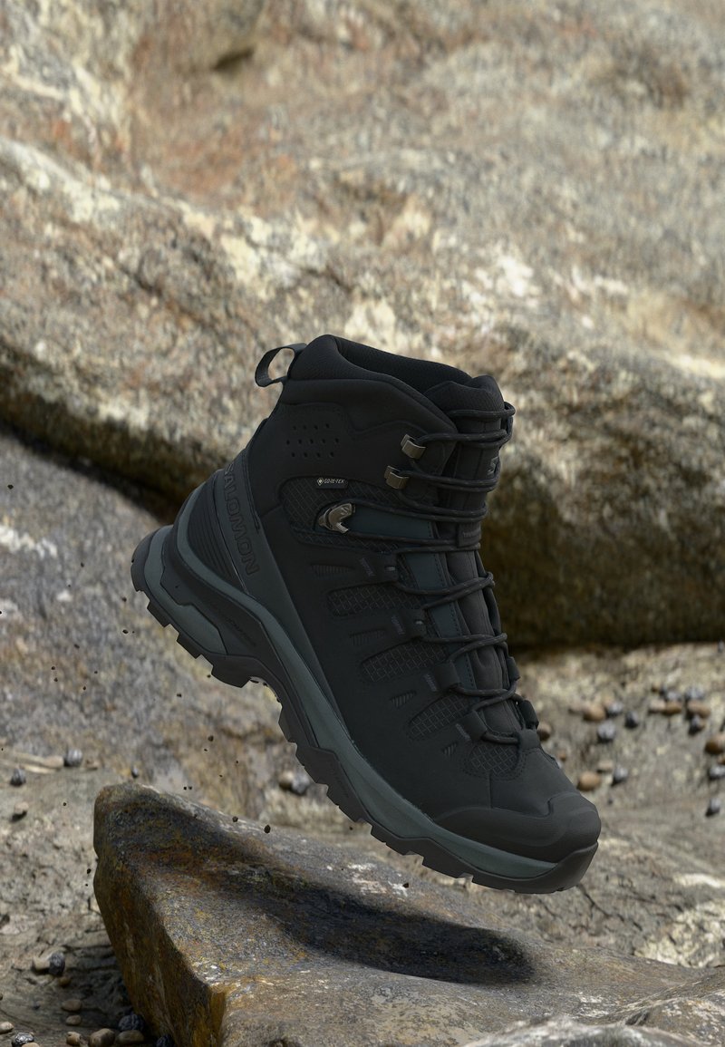 Μαύρο αναρριχητικό μποτάκι Salomon Gore-Tex κρεμασμένο στον αέρα πάνω από πετρώδη έδαφος, υπογραμμίζοντας την ανθεκτική σόλα και τον στιβαρό σχεδιασμό.
