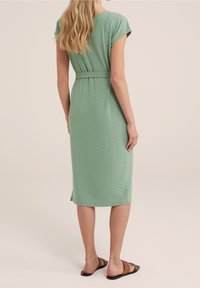 Vihreä polvipituinen mekko, jossa on lyhyet hihat, kuvioitu pinta ja vyötäröllä vyö. Yhdistetty ruskeisiin litteisiin sandaaleihin.