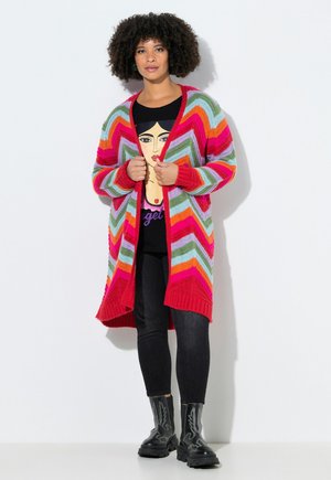 Nainen, jolla on kiharat hiukset, pukeutuneena moniväriseen siksak-kuvioiseen neuletakkiin, mustaan graafiseen t-paitaan, mustiin farkkuihin ja mustiin kuviollisiin saappaisiin, seisoo valkoista seinää vasten.