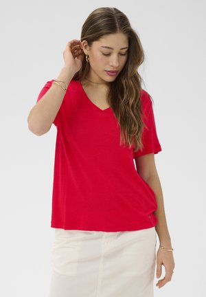 Junge Frau mit langen Haaren, die ein rotes V-Ausschnitt-T-Shirt, einen weißen Rock und goldenen Schmuck trägt, posiert mit einer Hand nahe am Ohr.