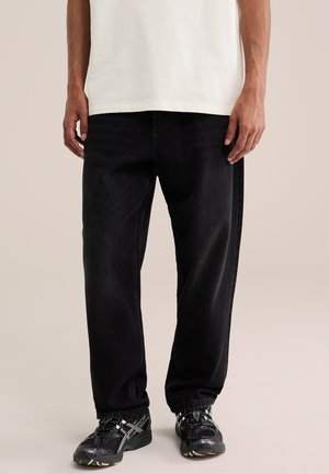 Homme portant un jean noir ample et des chaussures de sport noires, debout les mains détendues le long du corps devant un fond uni.