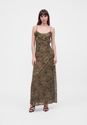 Woman with long brown hair wears a sleeveless leopard-print maxi dress with ruffle detail and black high-heeled sandals, standing against white background.
