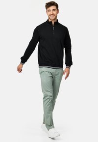 Svart zip-neck genser med ribbestrikkede mansjetter og kant, sammen med lysegrønne slim-fit bukser og hvite sneakers. Enkel tekstur, uten mønster.