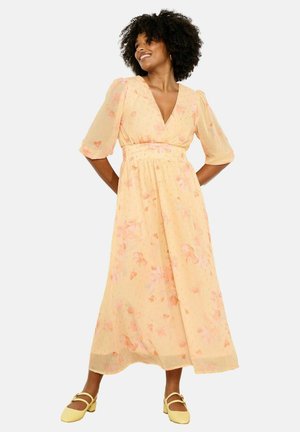 Naeratav naine lokkis juustega, kandes helekollast lillelist midi-kleitiga, millel on paugvarrukad, ning kollaseid Mary Jane tüüpi kingi, käed selja taga.