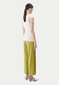 Kvinna med långt mörkt hår i en vit ärmlös topp, gula ribbade vida byxor, silverfärgade slingback-höga klackar, står mot en enkel bakgrund.