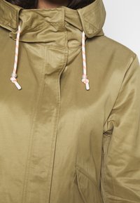 Khaki nepromokavá bunda s vysokým límcem a stahovacími šňůrkami. Disponuje hladkým, lesklým materiálem a předními kapsami, což podtrhuje minimalistický design.