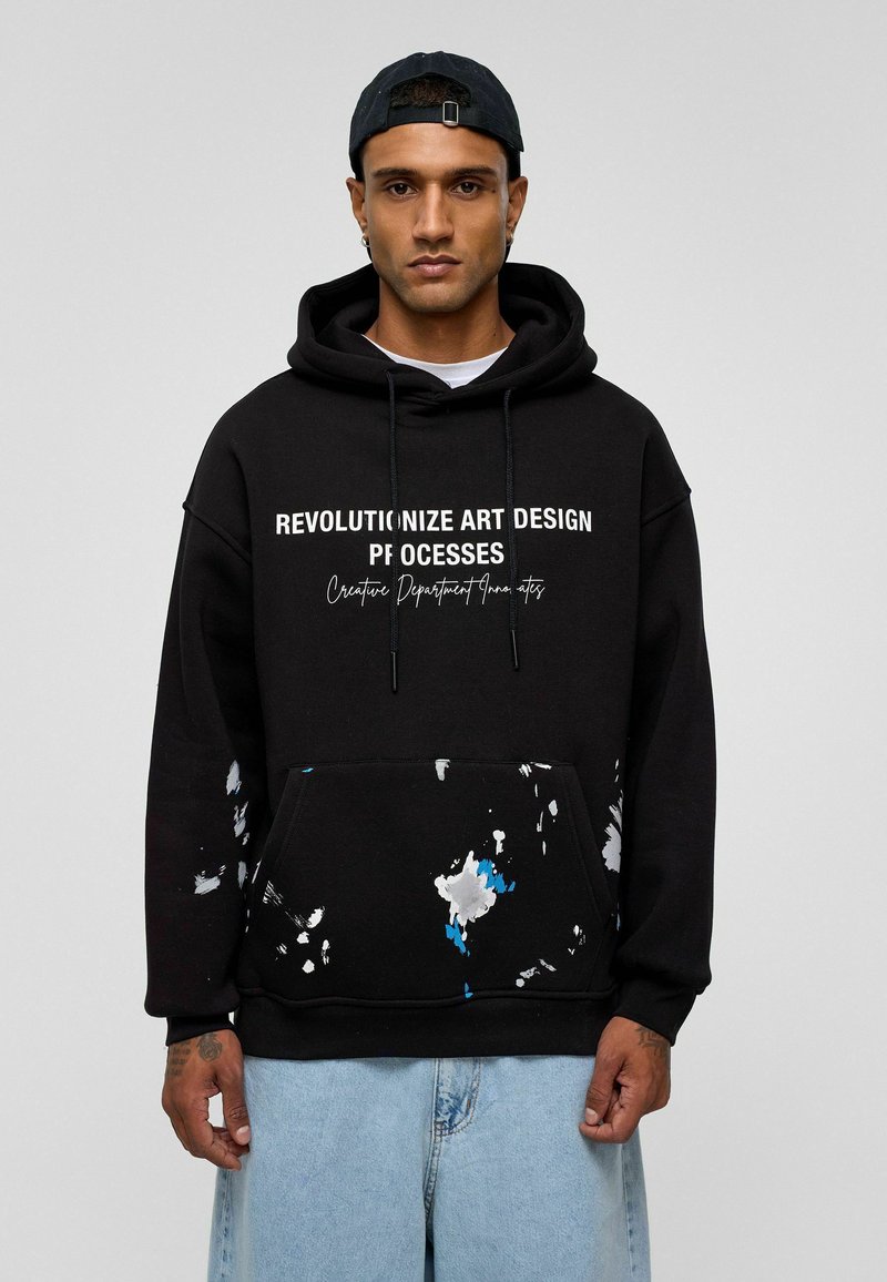 Zwarte hoodie met een voork pocket, voorzien van witte tekst en kleurrijke verfspetters in blauw en wit aan de onderzoom.