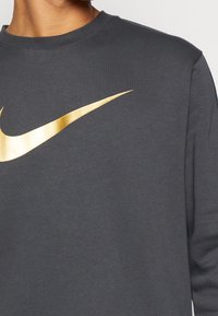 Mörkgrå sweatshirt i bomull med rund halsringning, prydd med en stor metallisk guldig Nike-logotyp framtill. Slät textur.