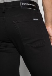 Närbild av svarta Calvin Klein-jeansfickor och midjeband på en person som bär en grå skjorta.