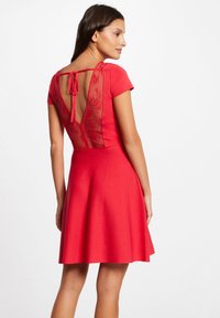 Rotes Kleid mit kurzen Ärmeln, tailliertem Oberteil und schwingendem Rock. Durchsichtiger Rückenbereich mit besticktem Design und einer Schleifenverschluss. Glatte Stoffstruktur.