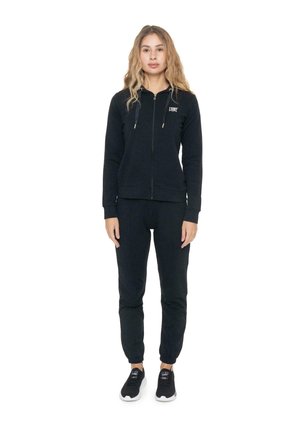 Una mujer luce una sudadera negra con capucha y cremallera, unos pantalones de chándal a juego y unas zapatillas negras sobre un fondo blanco liso.