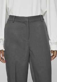 Pantalones grises a medida con una textura suave, que presentan un frente plano, pasadores para cinturón y bolsillos laterales, combinados con una blusa blanca de mangas largas.