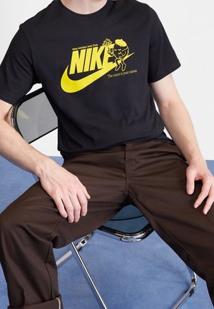 Osoba ubrana w czarną koszulkę Nike z żółtym logo i grafiką, siedząca na czarnym metalowym składanym krześle na niebieskiej podłodze.