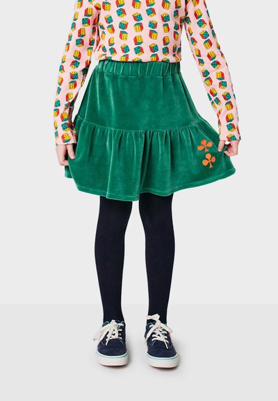Jupe émeraude en velours côtelé, à volants, dotée d'une taille élastique et ornée d'un détail floral orange, assortie de collants noirs et de baskets bleues.