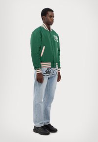 Grønn sateng bomberjakke med stripete mansjetter, med et bokstavlapp. Brukt over lyseblå jeans med svart grafisk design og svarte sko.