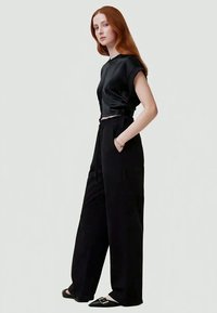  Schwarzes Satin-Oberteil mit kurzen Ärmeln, kombiniert mit hochgeschnittenen schwarzen Hosen und flachen schwarzen Schuhen mit quadratischer Schnalle.