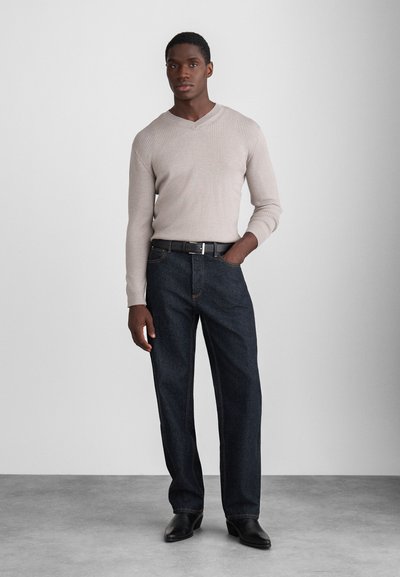 Bežani rebrasti pleteni pulover s V-izrezom, upotpunjen tamnoplavim trapericama s vidljivim šavovima i crnim remenom, te crnim cipelama.