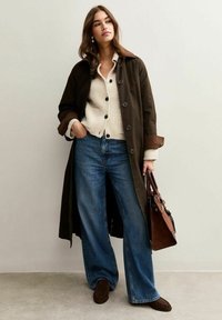 Manteau long marron avec des détails de boutons sur un cardigan crème, associé à un jean bleu à jambes larges et des mocassins marron. Porte un sac à main marron.