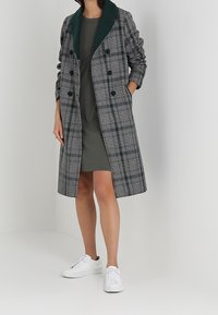 Frau in einem karierten langen Mantel mit grünem Kragen über einem grauen Kleid und weißen Sneakers, die mit den Händen in den Manteltaschen steht.
