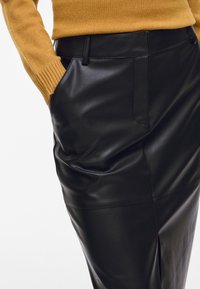 Κοντινό πλάνο ενός ατόμου που φοράει ένα μουσταρδί πουλόβερ και μαύρη δερμάτινη φούστα με ψηλή μέση, με το ένα χέρι στην τσέπη της φούστάς.