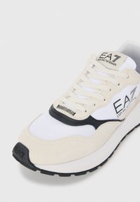 Beige and white sneaker featuring a mesh upper, suede accents, black detailing, and a logo on the tongue. Black outsole with textured design.