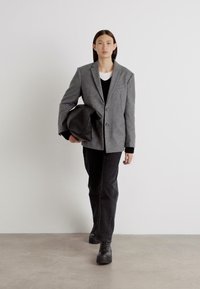 Blazer gris avec deux boutons, pull en laine noir en dessous, pantalon noir et chaussures noires. Le modèle tient un grand sac noir.
