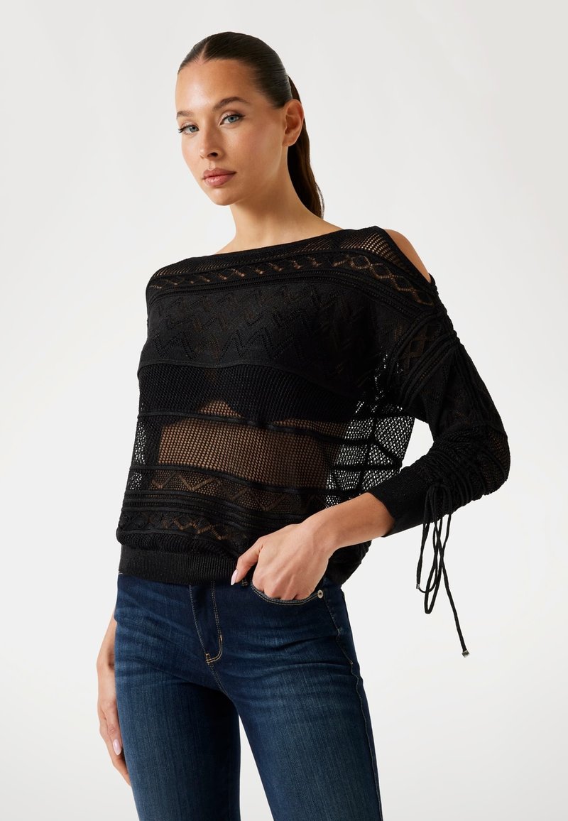 Zwarte gebreide trui met open schouders, gestructureerde patronen en lange mouwen. Gecombineerd met een donkerblauwe slim fit jeans.