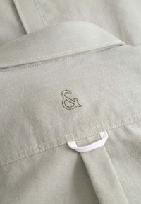Helles beige Gewebe mit glatter Textur, das ein dezentes "&"-Logo in grün eingestickt hat. Enthält eine weiße Schlaufe und saubere Nähdetails.