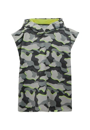 Mädchen mit lockigem Haar, das eine grau-gelbe Camouflage-ärmellose Kapuzenjacke und passende Shorts trägt, steht barfuß auf einfarbigem Hintergrund.