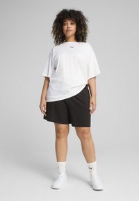 Balta per didelį T-shirt su mažu juodu logotipu, juodos šortai ir baltos sportinės kedai. Tekstūra lygi; audinys atrodo pralaidus orui.