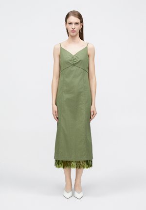 Ženska v olivno zelenih brezrokavnih midi obleki z nabrano oprsno preklopno in robom z bleščečimi resicami, obuta v bele čevlje z ostro konico.