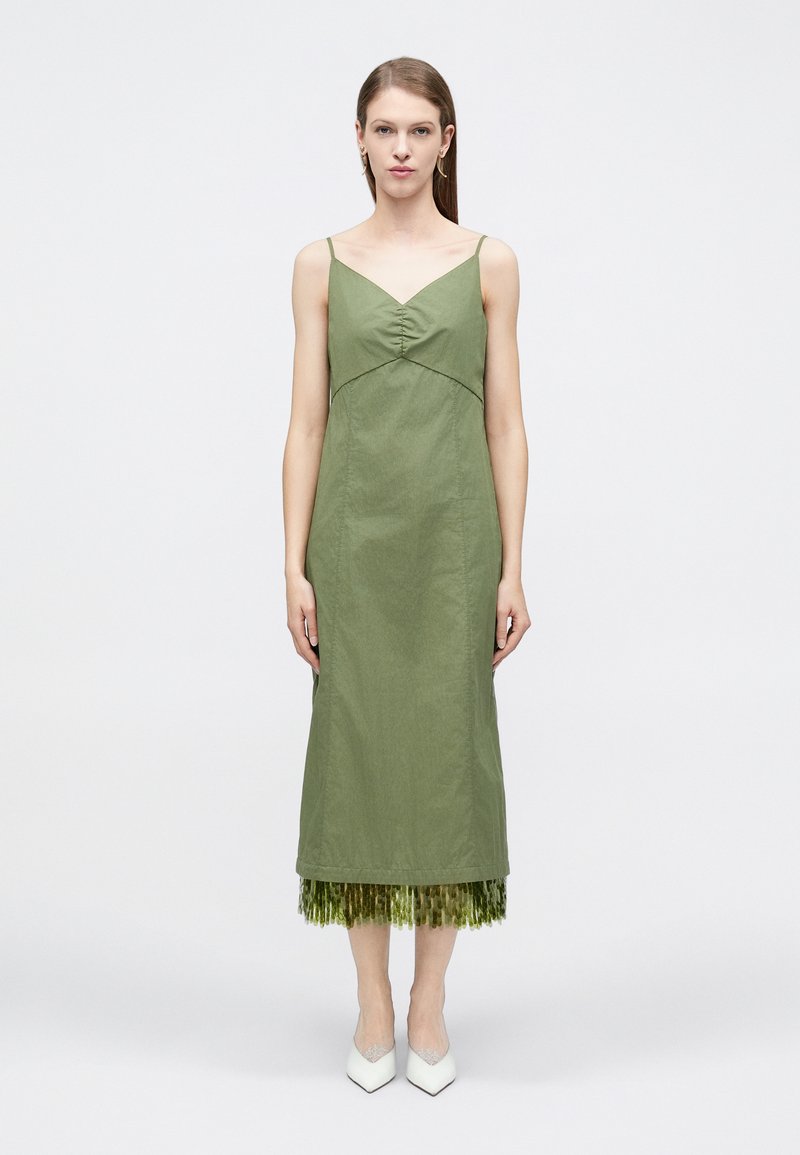 Γυναίκα που φοράει μια αμάνικη μεσαίου μήκους ελιάς πράσινη φόρεμα με σούρες στο μπούστο και φράντζα με παγιέτες στο τελείωμα, συνδυασμένη με λευκές μυτερές γόβες.