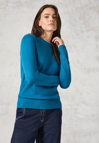 Pull en tricot bleu avec un col rond et des poignets côtelés. Texture lisse et coupe ajustée, associé à un jean noir. Design subtil, sans motifs.