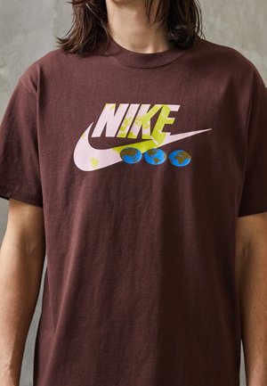Henkilö pukeutunut ruskeaan t-paitaan, jossa iso vaaleanpunainen Nike-logo ja kolme pientä sinivihreää maapallon kuvaa logon alapuolella.