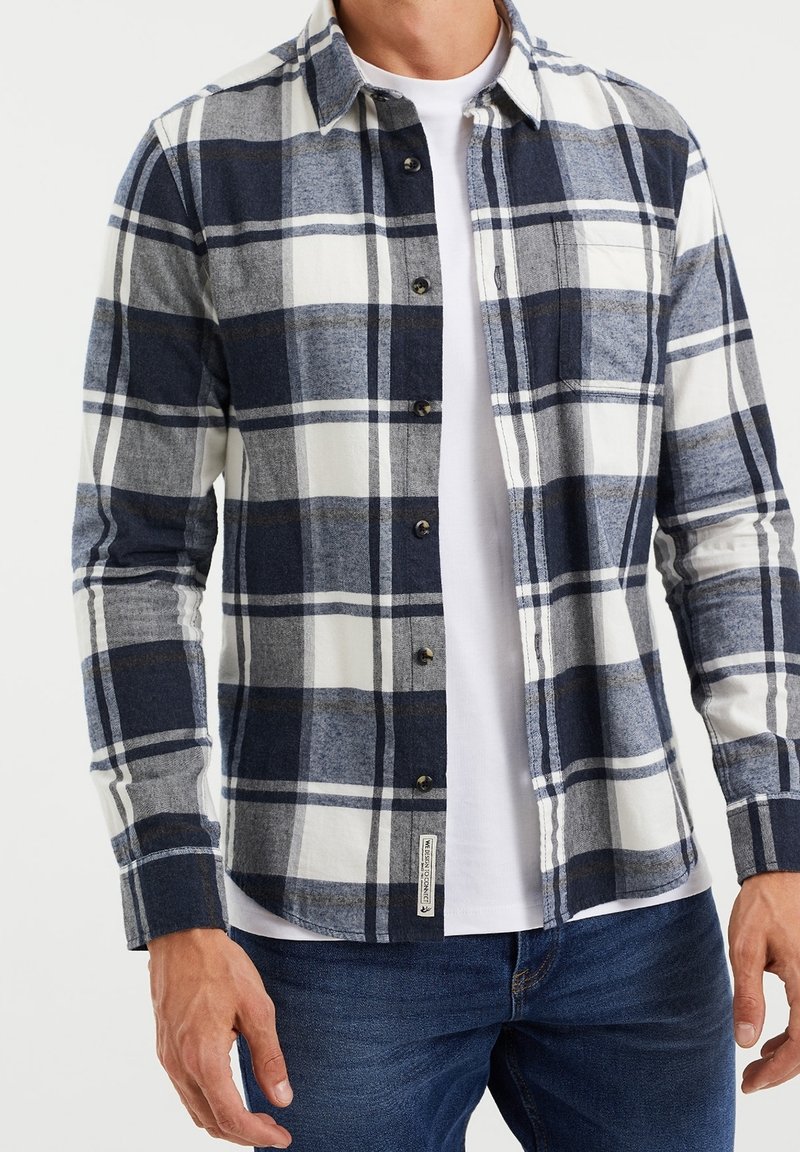 Herren Flanellhemd in Navy-, Grau- und Weißkaros mit Knopfverschluss, spitzem Kragen und langen Ärmeln. Getragen über einem weißen T-Shirt.