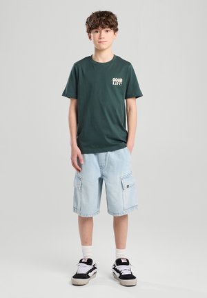 Grünes Baumwoll-T-Shirt mit Logo, kombiniert mit leichten blauen Cargoshorts mit Taschen. Schwarze und weiße Sneaker runden das Outfit ab.