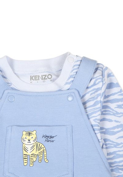 Salopette bleu clair avec une poche avant ornée d'un graphique de tigre, associée à une chemise blanche affichant un motif de zèbre bleu. Tissu doux, design décontracté.