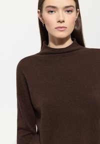 Eine Frau mit glattem braunem Haar trägt goldene Creolen und einen dunkelbraunen Rollkragenpullover vor einem schlichten hellen Hintergrund.