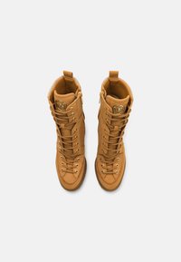 Bottes montantes à lacets couleur beige avec fermetures éclair latérales et un emblème circulaire de la marque sur la languette, vues de dessus sur fond blanc.