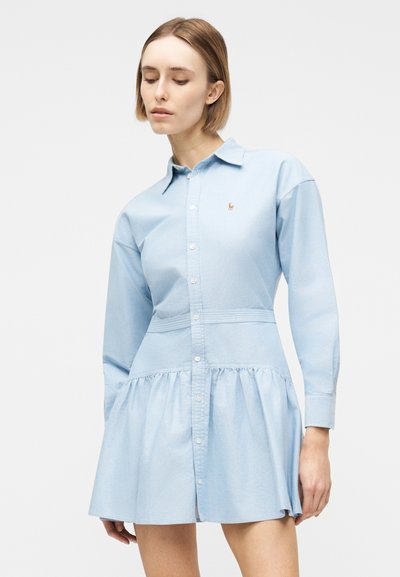 Νεαρή γυναίκα με κοντά μαλλιά που φορά ένα γαλάζιο φόρεμα με μακριά μανίκια, κουμπιά και ένα μικρό κεντημένο λογότυπο στο στήθος.