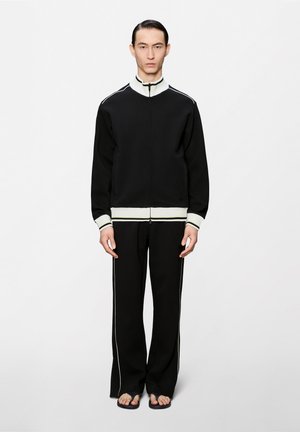 Άνδρας μοντέλο με μαύρο μπουφάν φόρμας με φερμουάρ και λευκή λεπτομέρεια, μαύρο παντελόνι με λευκές ρίγες στα πλαϊνά και μαύρα σαγιονάρια, στέκεται μπροστά σε λευκό φόντο.