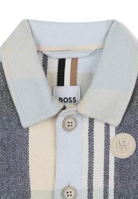 Camicia con colletto a righe nei toni del blu chiaro, crema e blu scuro. Due bottoni e una patch con logo sul petto.
