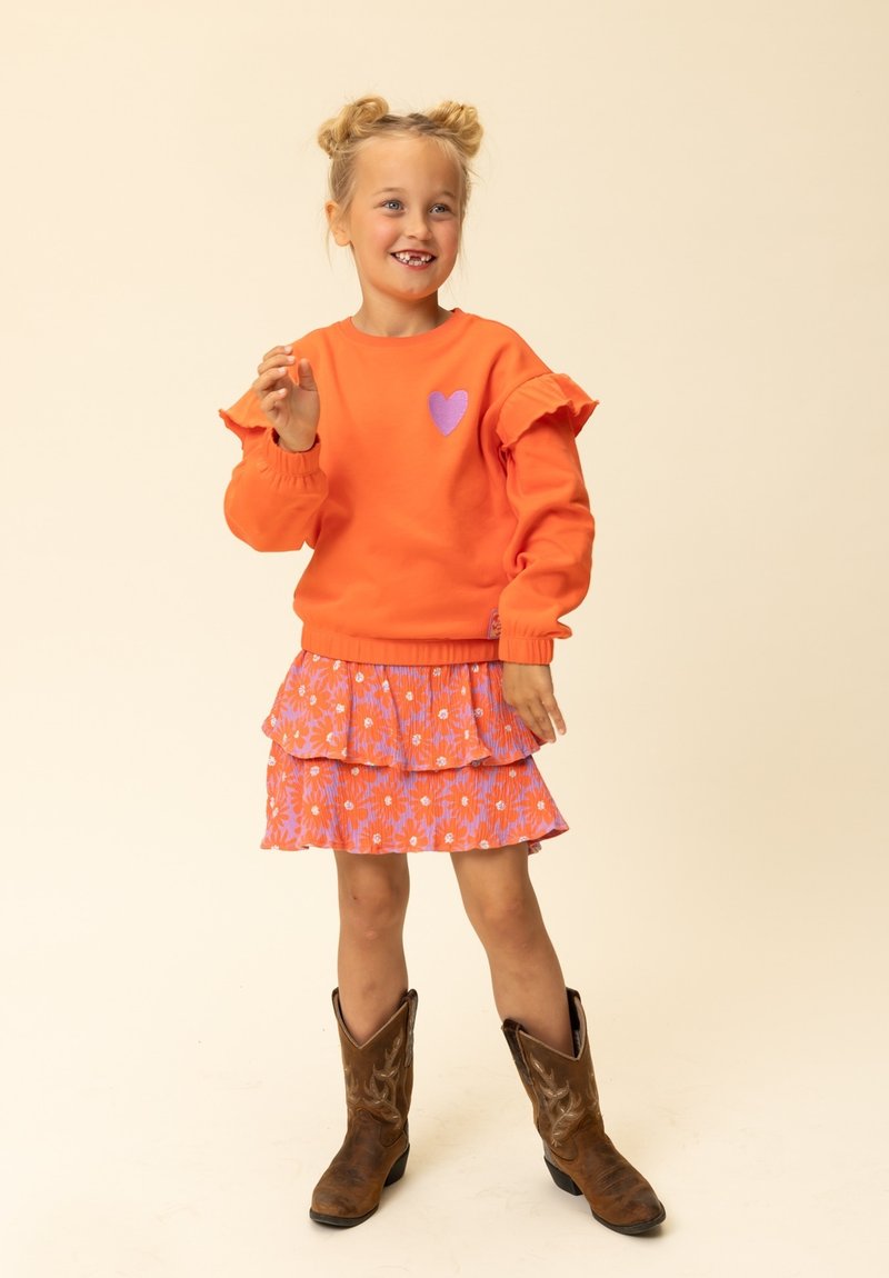 Oranje sweatshirts met ruches aan de mouwen en een paarse hartapplicatie, gecombineerd met een roze bloemenrok in lagen en bruine cowboylaarzen.