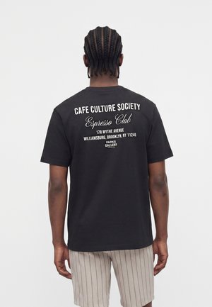 Mand med fletninger, iført en sort t-shirt med hvid tekst om Cafe Culture Society og beige, stribede shorts, set bagfra.