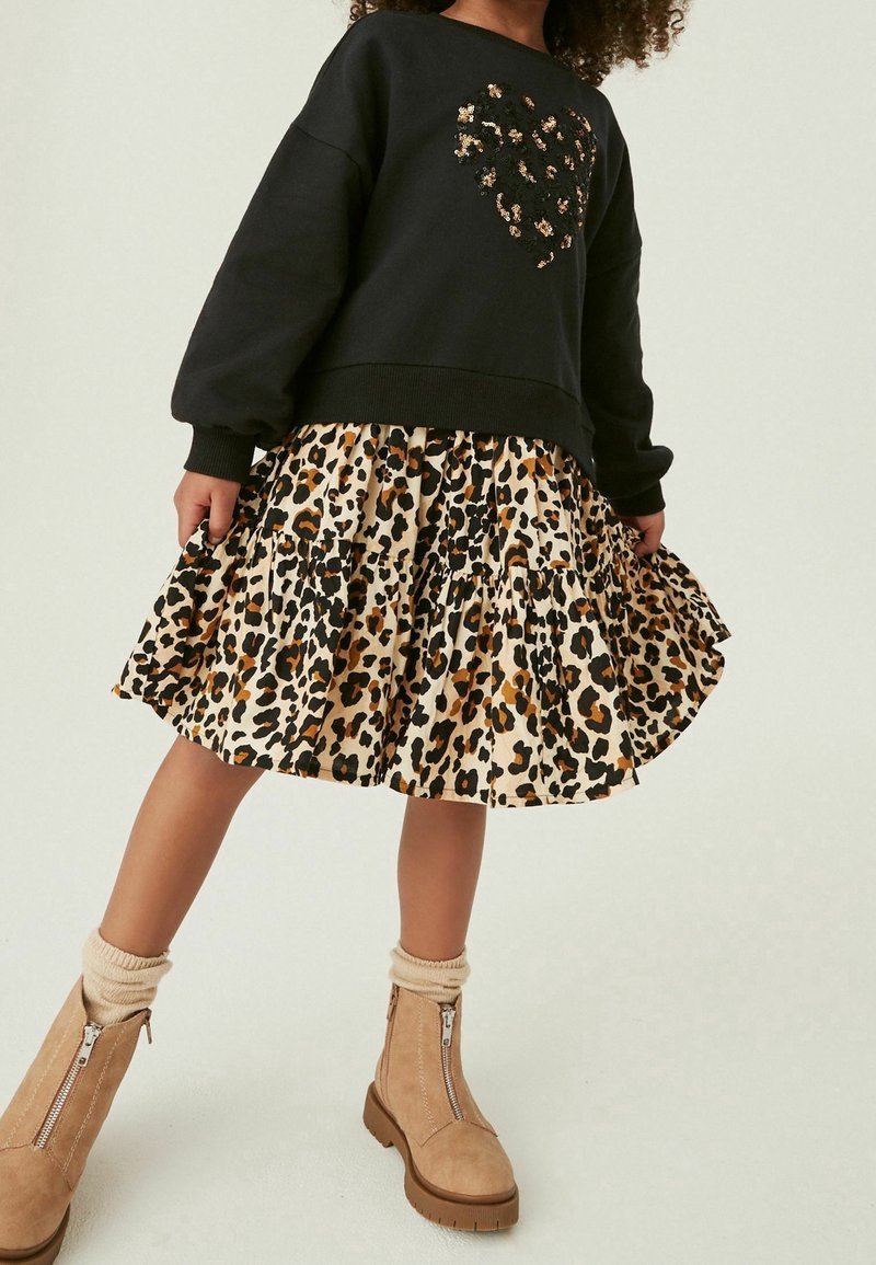 Sweat-shirt noir avec un motif de cœur, associé à une jupe en tiered imprimé léopard. Le mannequin porte des bottines beiges avec des zips et des chaussettes crème.