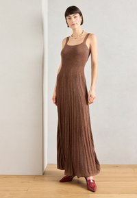 Braunes, geripptes Maxi-Kleid mit quadratischem Ausschnitt und breiten Trägern. Es besitzt einen fließenden, gefältelten Rock. Kombiniert mit roten, spitzen Schuhen.