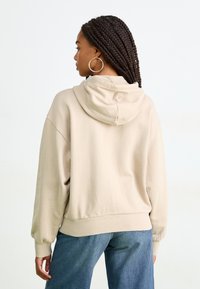 Beige oversized hettegenser med snor i hetta, ribbede ermer og kant, kombinert med blå avslappede jeans. Mykt stoff og minimalistisk design.