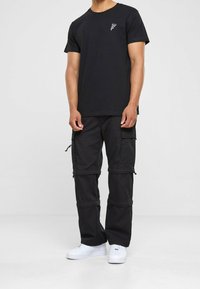Černé tričko s malou grafikou na levé hrudi, spojené s černými cargo kalhotami s kapsami a zipy. Oblečení doplňují bílé tenisky.
