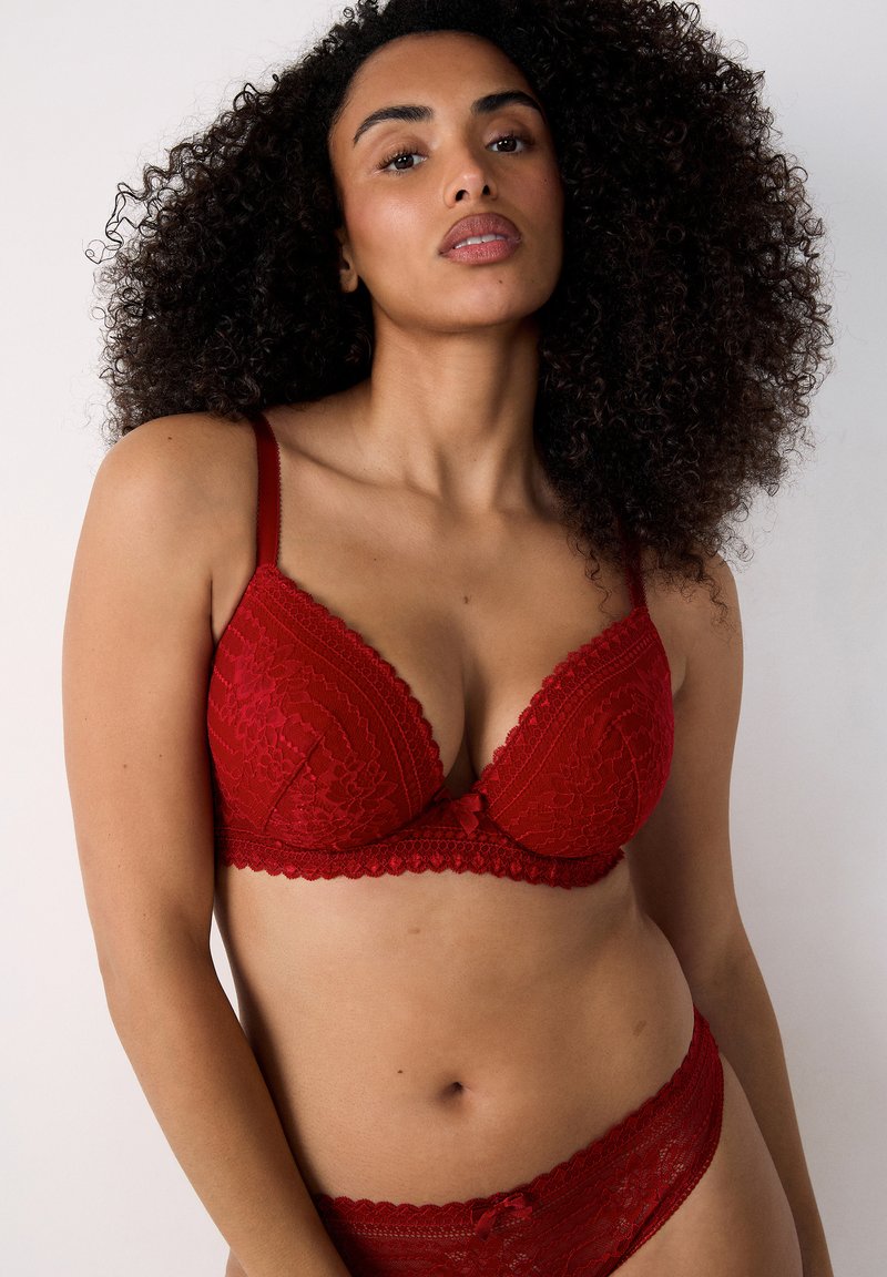 Czerwona koronkowa bielizna z głębokim dekoltem, ozdobnymi falbany na brzegach oraz regulowanymi ramiączkami. Posiada centralny detal w postaci kokardy oraz pasujące koronkowe majtki.