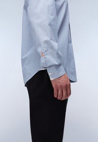 Blauw-witte gestreepte lange mouwen shirt met knoopmanchetten, voorzien van een klein oranje logo op de manchette, gedragen met donkere broek.